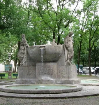 Nornenbrunnen stone fountain with three sculpted figures around a central basin and water flowing into a lower pool.