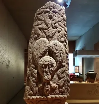 Pfalzfeld Obelisk with a carved human face and swirling motifs in a museum display