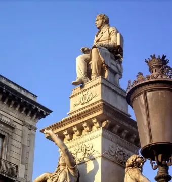 Monument to Vincenzo Bellini with a seated white marble statue on a decorated pedestal, lit by warm sunlight beside a streetlamp.