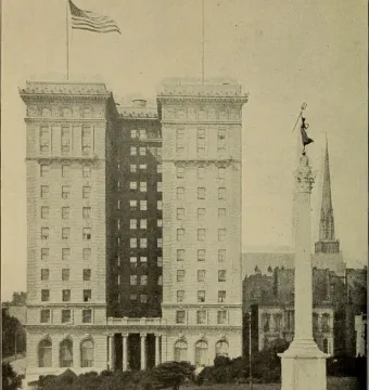 Dewey Monument rising from a circular basin in a park with buildings and a church steeple behind it.
