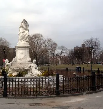 The Lorelei Fountain, a light-colored sculpted stone monument, stands behind a black metal fence in a park with bare trees and city buildings.