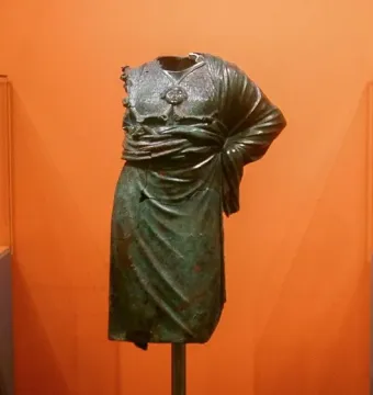 Headless, armless Minerva d\'Arezzo torso on a pole, displayed between museum cases with a detached arm fragment and a helmeted head sculpture.