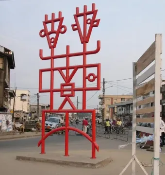 Njé Mo Yé, a bright red abstract sculpture with geometric lines and two stylized figures, stands on a concrete base beside a busy street.