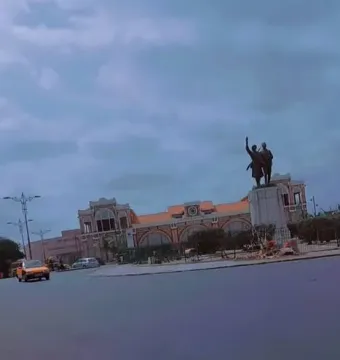 The Demba et Dupont statue stands on a light-colored pedestal near a plaza, with a large building and cloudy sky in the background.