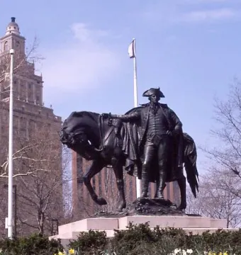 George Washington bronze sculpture beside a harnessed horse on a stone pedestal, with flowers in front and a towered building behind.