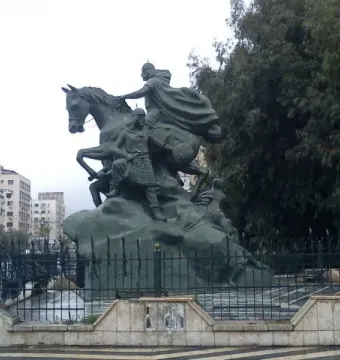 Statue of Saladin, a dark equestrian monument with a cloaked rider on a rearing horse and figures at the base, in an urban square.