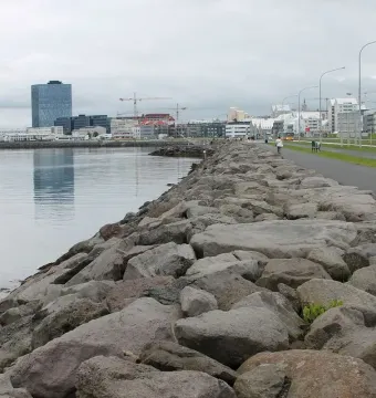 Rocky embankment beside calm water and a paved path with Reykjavik buildings in the distance.