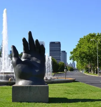 Dark hand sculpture with fingers pointing upward on a stone pedestal beside a fountain and lawn.