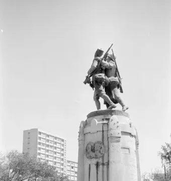 Demba et Dupont monument with two armed figures on a tall stone pedestal in a park-like square.