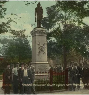 Firemen\'s Monument with a stone statue of a rescuer holding a child in a park, with men gathered nearby behind a red fence.