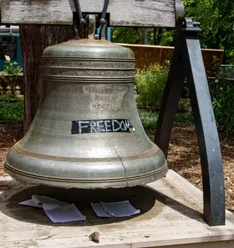 The Liberty Bell with a visible crack and the word “FREEDOM” taped over part of it, resting on a wooden platform with papers below.