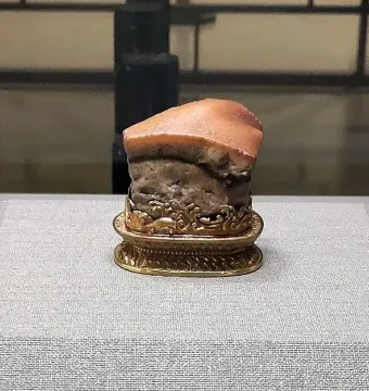 Meat-shaped Stone displayed on an ornate golden stand, resembling a piece of cooked pork belly with a reddish-brown top and mottled grey-brown body.
