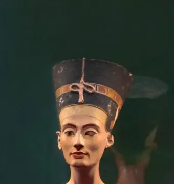 Bust of Nefertiti with a tall blue and gold crown, painted face, and patterned collar against a dark green background.