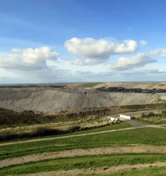Northumberlandia’s vast sculpted earthworks rise beyond grassy slopes and dirt paths under a partly cloudy sky.
