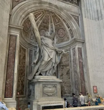 Saint Andrew sculpture standing in an ornate alcove inside St. Peter\'s Basilica, with a carved pedestal and visitors below.