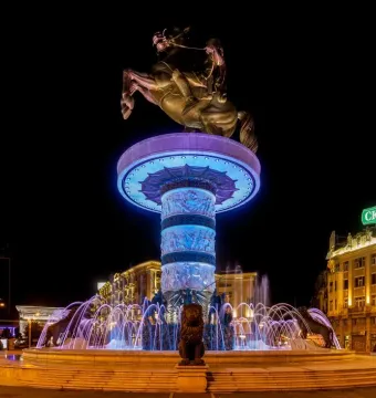The Warrior on a Horse statue lit in blue and purple above a brightly illuminated fountain with lion sculptures at night.