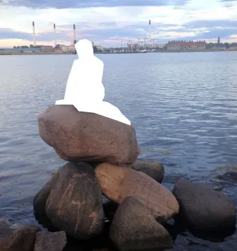 White silhouette of the Little Mermaid seated on rocks by calm water with a distant city skyline