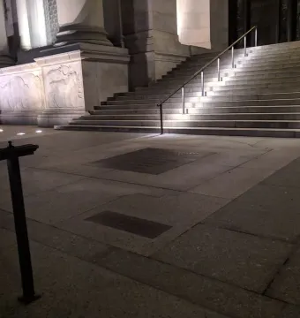 Nighttime view of the American Museum of Natural History plaza with illuminated stone reliefs and inscribed plaques in the foreground.