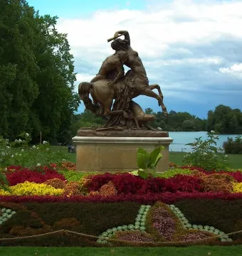 Centauress and Faun bronze sculpture on a stone pedestal amid flower beds by the water.