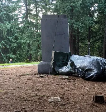Toppled Harvey W. Scott statue lying beside a tall stone pedestal among trees.