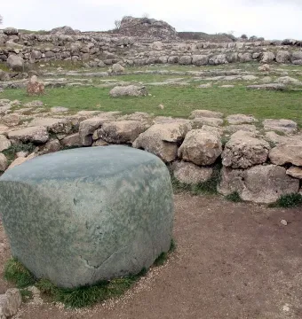 A large green stone block in front of Hattusa archaeological ruins with grassy foundations under an overcast sky.