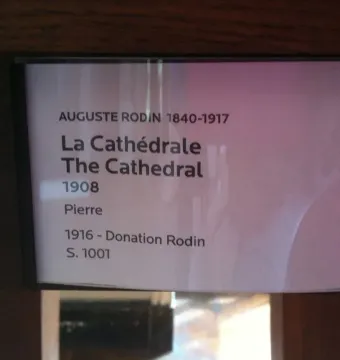 Museum information plaque for Auguste Rodin’s La Cathédrale, with a blurry pink reflection at the right edge.