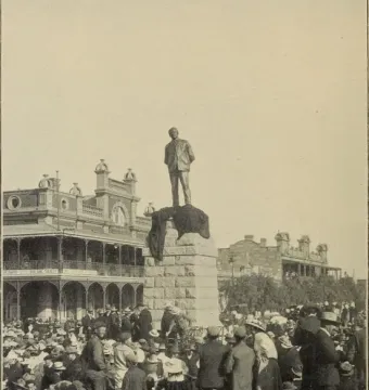The statue of Cecil Rhodes rises on a tall stone pedestal in a crowded public square, with historic buildings in the background.