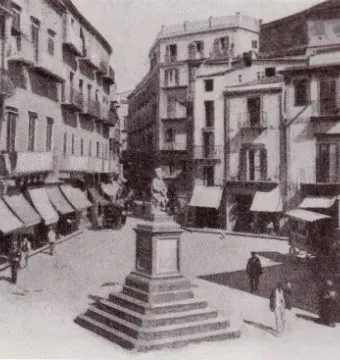 The Fountain of the Genius of Fieravecchia in a town square, with a small seated figure on a tiered pedestal surrounded by buildings and storefronts.