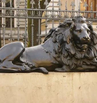 Guarding Lion statue of a resting lion on a light-colored plinth in front of an ornate metal fence and building facade.