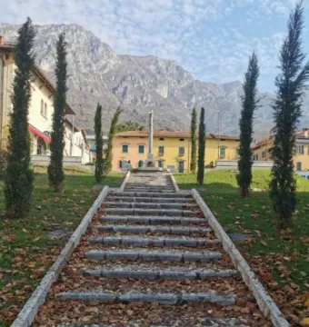 Stone stairs covered with autumn leaves lead up to the Lighthouse of Lierna on a grassy slope, framed by cypress trees and mountains beyond.