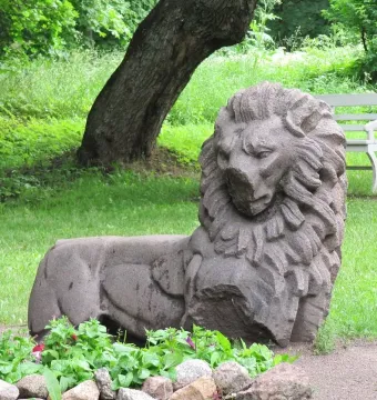 Weathered stone lion sculpture resting on the ground in Mon Repos park, with a tree trunk, bench, and flower bed nearby.