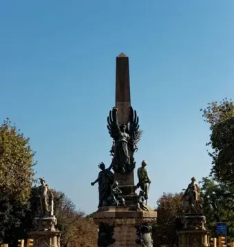 Rius i Taulet Monument with dark bronze figures at its base in Barcelona, with pedestrians and trees nearby.