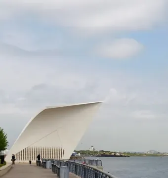 The Staten Island 9/11 Memorial postcard-shaped white monument stands on a waterfront boardwalk beside metal railings and water.