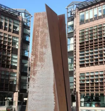 Fulcrum, a tall rust-colored steel sculpture standing between brick and glass buildings in an urban plaza.