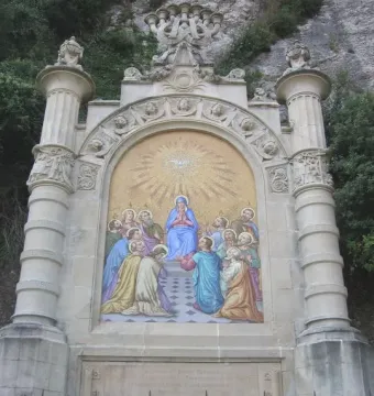 Monumental Rosary of Montserrat with a colorful Pentecost mosaic in a stone arch, seen behind a black metal fence.