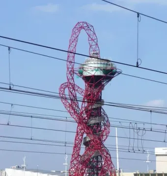 Orbit Tower with a red twisting lattice structure and circular platform near the top, seen behind overhead power lines and railway tracks.
