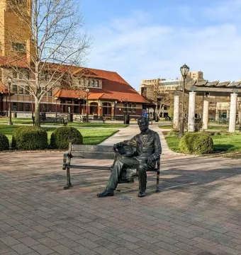 Seated Lincoln sculpture on a bench in a brick-paved public square with landscaped shrubs and buildings nearby.