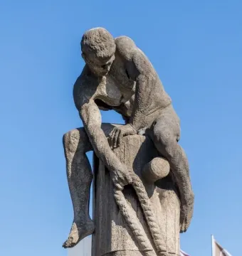 Tauzieher, a weathered stone sculpture of a muscular man pulling a thick rope around a stone post.
