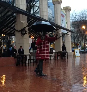 Allow Me sculpture of a figure in a checkered coat holding an open umbrella on a wet brick plaza.