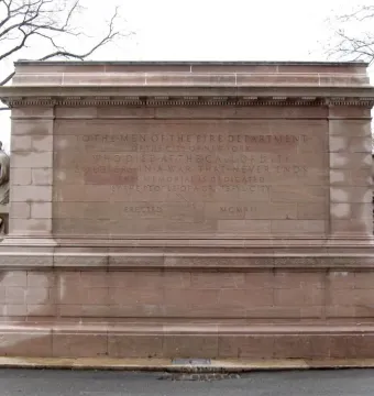 Firemen\'s Memorial stone monument with carved inscriptions and sculpted figures beside bare trees.