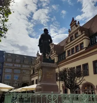 Goethe Monument seen from behind on a stone pedestal with inscriptions and a framed bust, surrounded by a green fence in Leipzig.