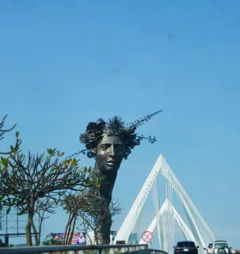 Reminiscencia, a dark metallic bust of a woman with a sculpted floral crown, stands outdoors against a blue sky.