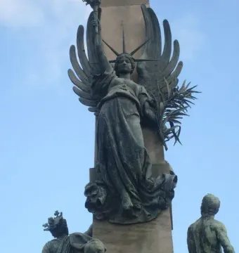 Close-up of the Rius i Taulet Monument showing a winged female figure holding a torch and frond, with smaller sculpted figures below.