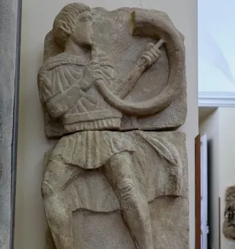 Broken stone relief of the Acrobat from Osuna showing a male figure playing a horn.