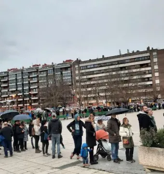 A large crowd gathers on a paved plaza under a cloudy sky, with umbrellas open and apartment buildings in the background.