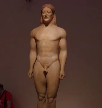 Kroisos Kouros, a nude male Greek statue with braided hair standing on a base in a museum setting.