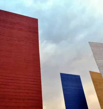Torres de Satélite geometric towers in red, blue, gray, and gold against a cloudy sky.