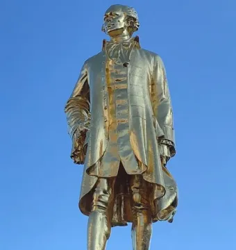 Golden statue of Alexander Hamilton in historical attire on a dark pedestal against a blue sky.