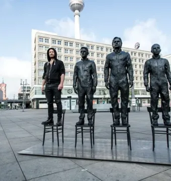 Anything To Say? sculpture in Berlin, with three bronze-like statues and a live person standing on chairs in a city plaza.