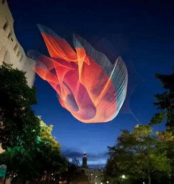 An enormous colorful textile installation floats above a city street at dusk.